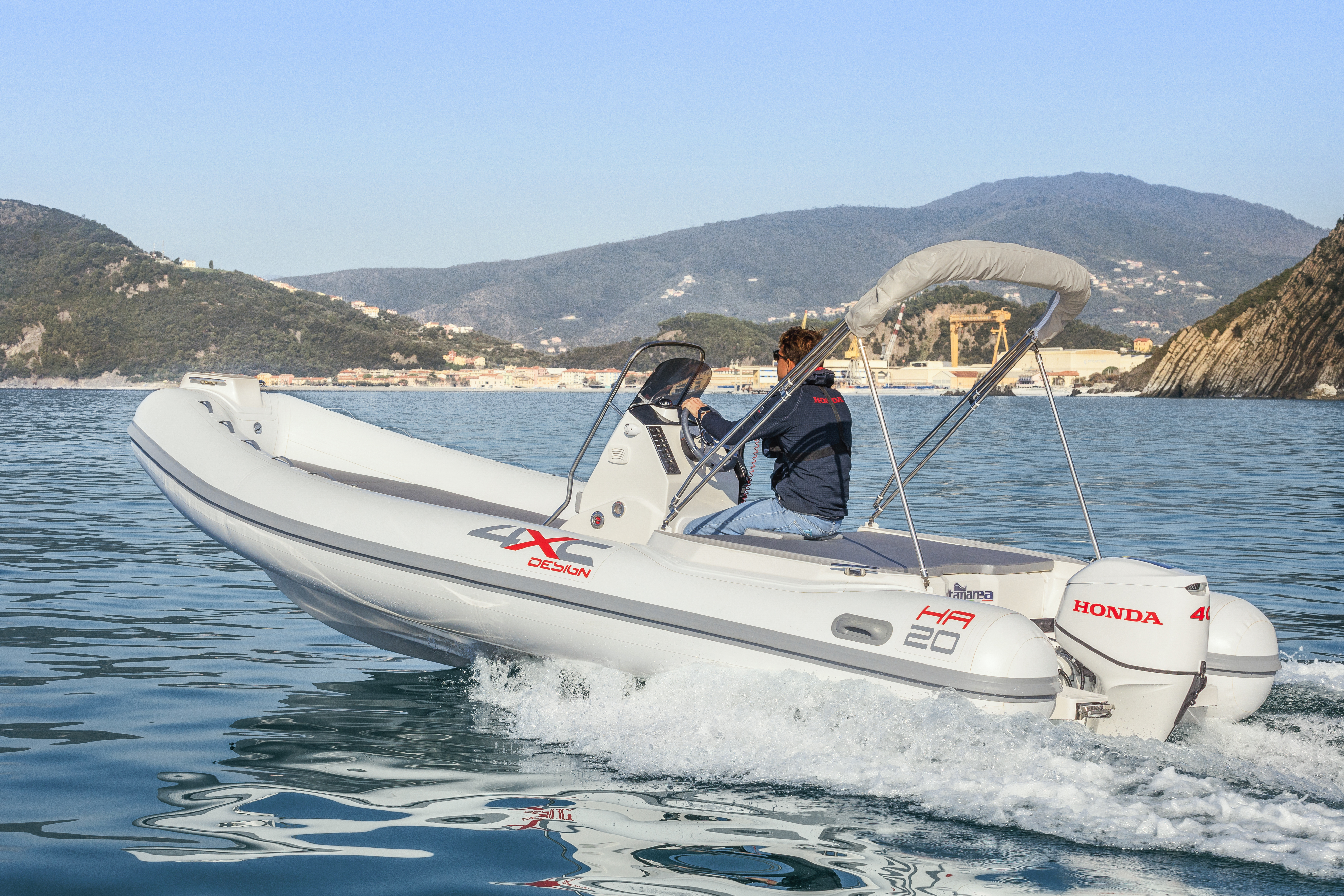 A white inflatable boat with a Honda outboard motor cruises on calm blue waters near a mountainous coastline. A person is seated at the helm, steering the boat.